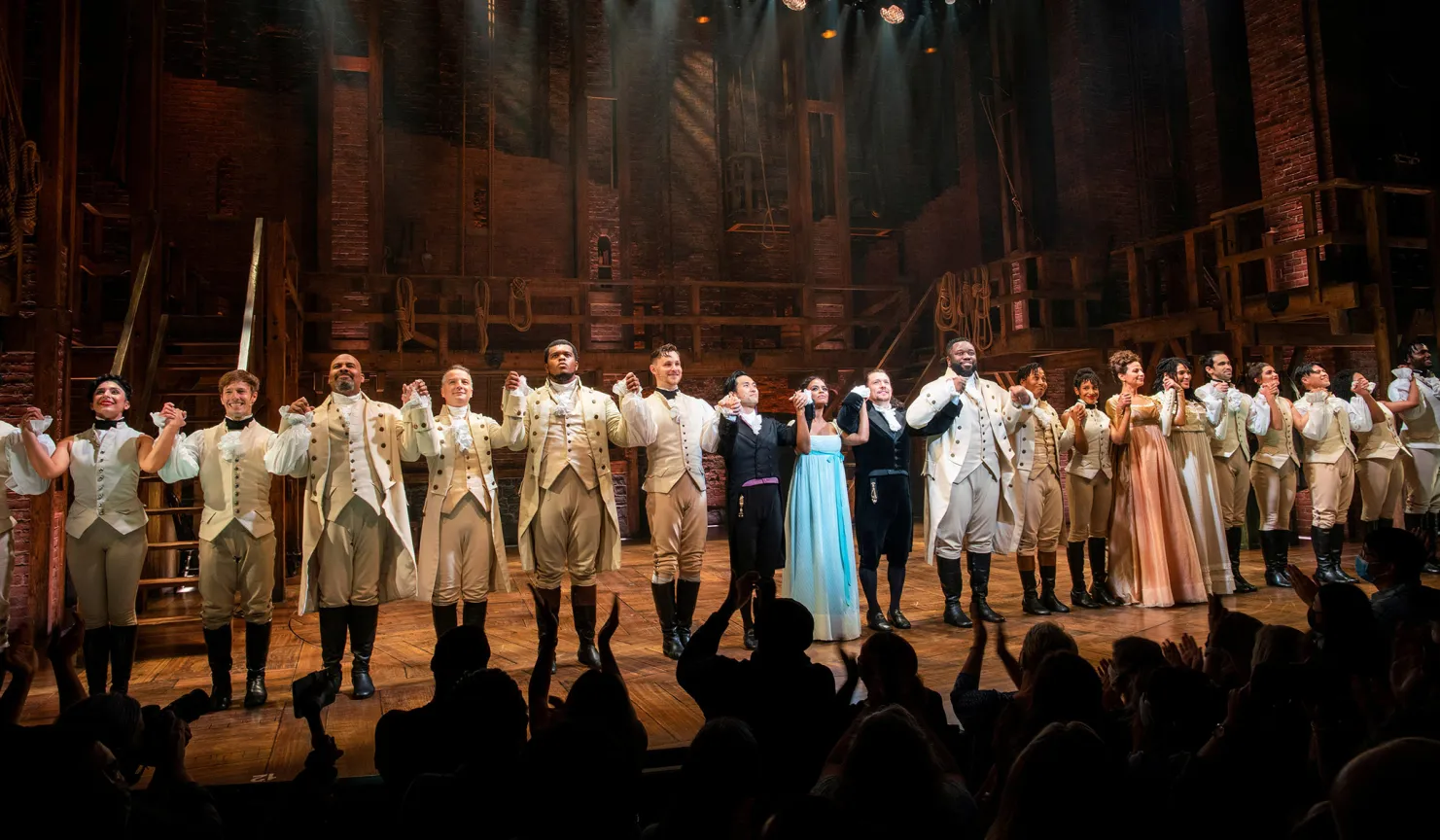 A Broadway theater during curtain call, with the cast lined up onstage under bright lights while audience members applaud, including patrons standing at the rear of the house