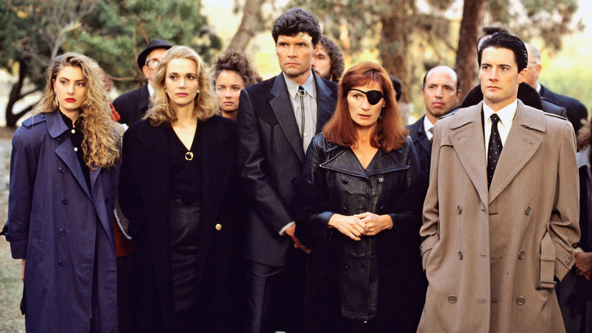 A candid behind-the-scenes photograph of a TV drama ensemble cast standing on a soundstage between takes while crew members adjust lighting