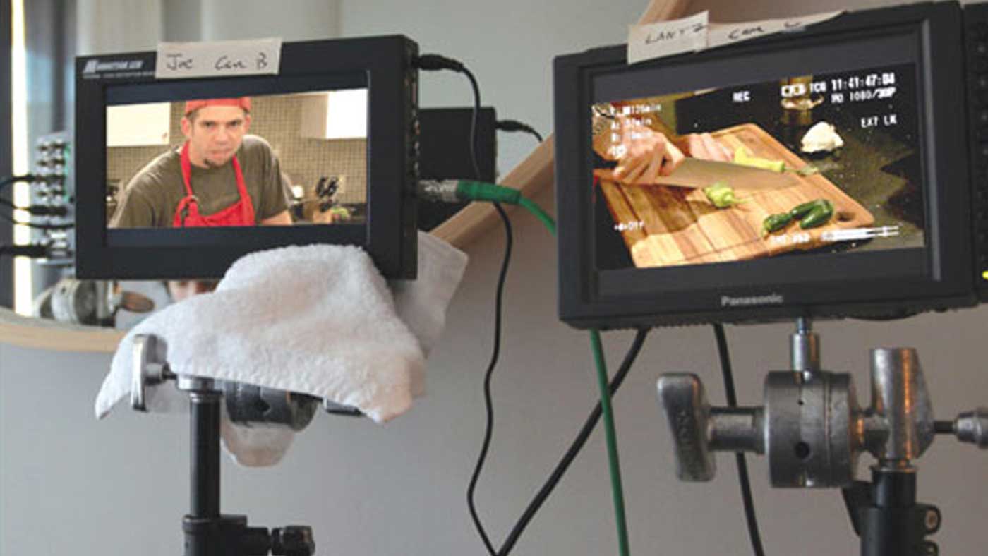 A casting director and a director seated near a video monitor on set, reviewing a take while crew members stand quietly behind them