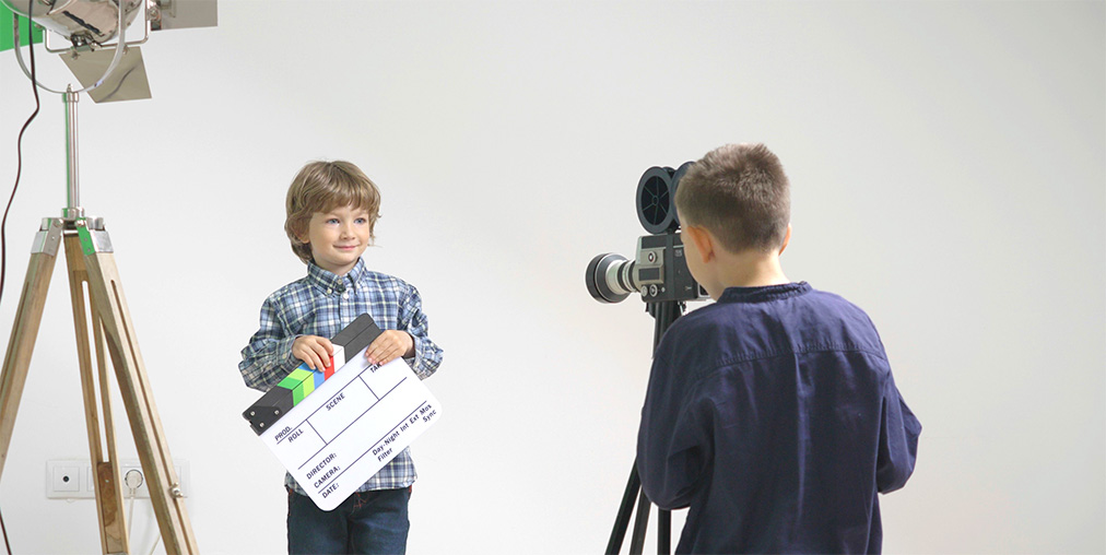 A child performer in wardrobe standing beside a parent or guardian near a film set holding area, with crew members working in the background