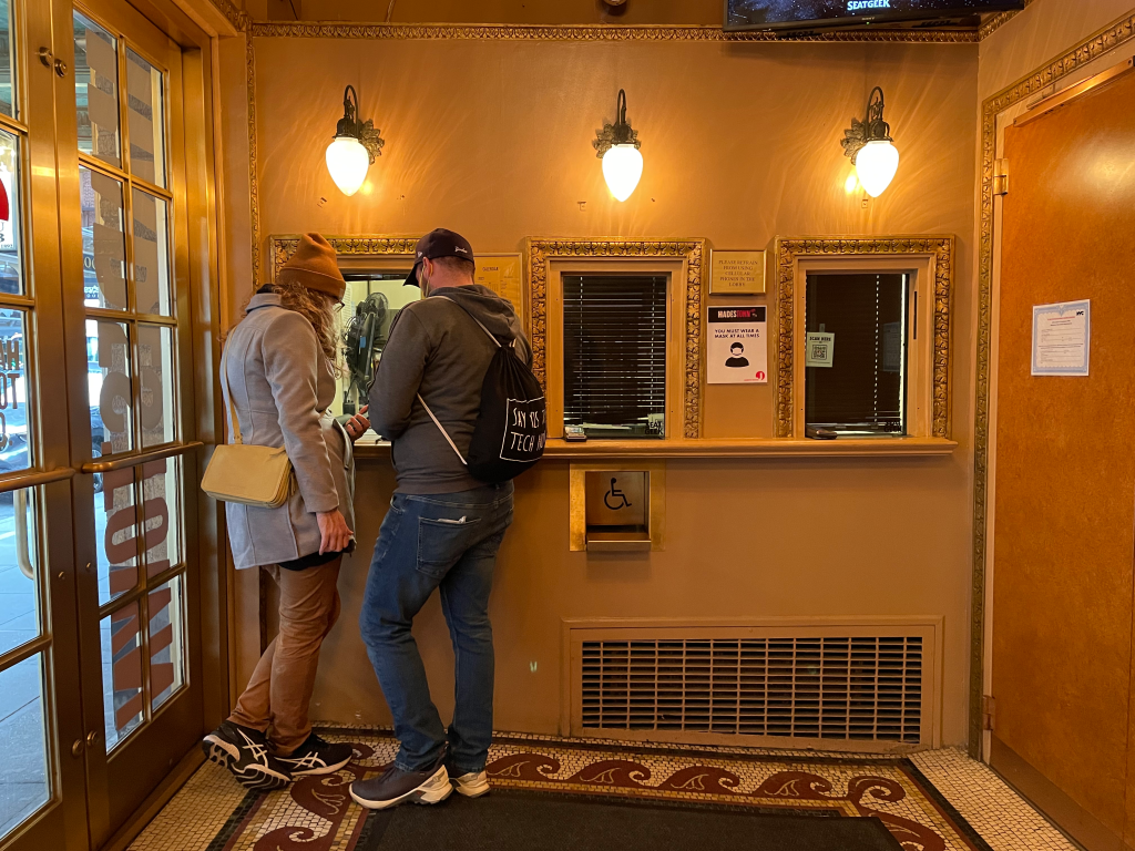 A close view of a Broadway theater box office window with posted performance times and a small line of patrons waiting