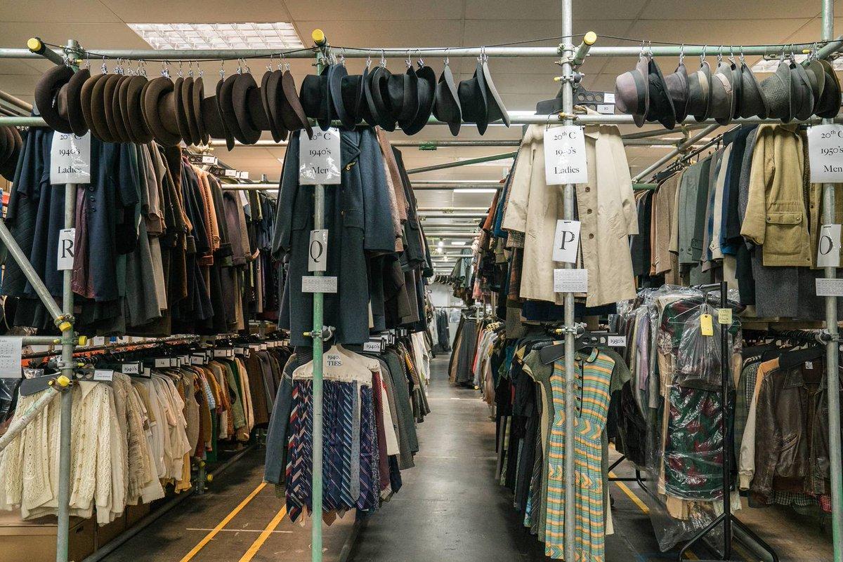 A costumer stands beside rolling clothing racks packed with labeled garments and hangers in a studio wardrobe area, overhead fluorescent lighting