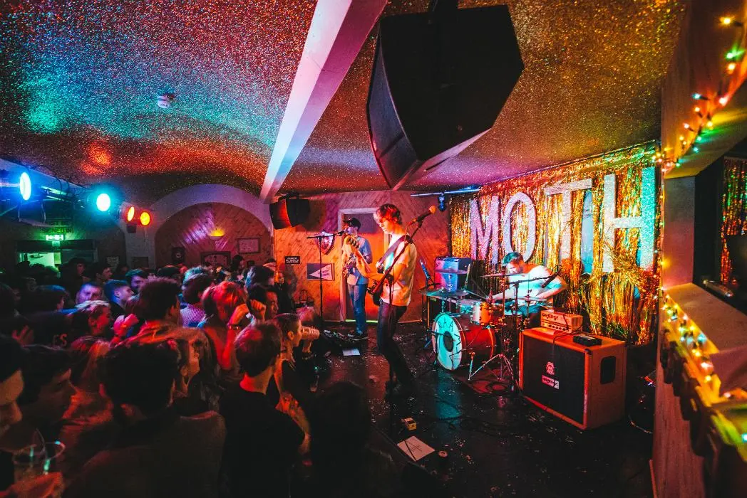 A crowded small music club with a standing-room floor facing a low stage, colorful stage lights reflecting on the audience