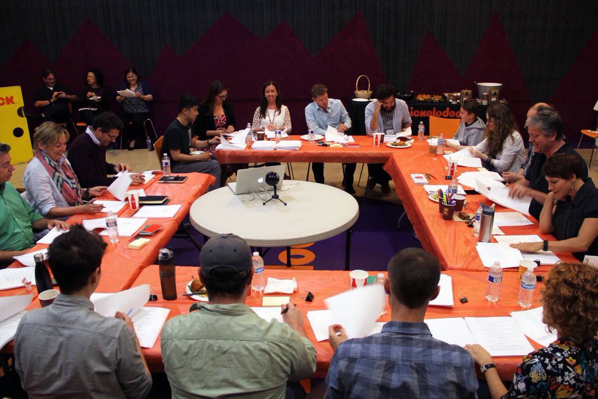 A director and two producers seated near the end of a conference table as actors read from stapled scripts, with water bottles and highlighters scattered across the table
