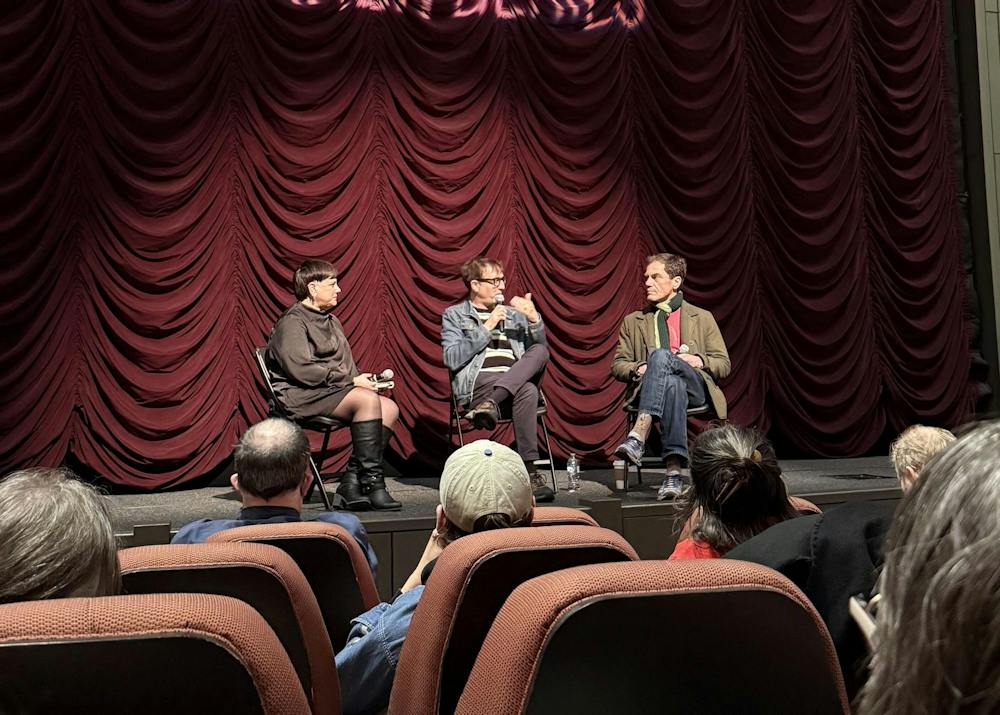 A director seated on a stage in front of a movie screen holding a microphone while audience members sit in a dim theater