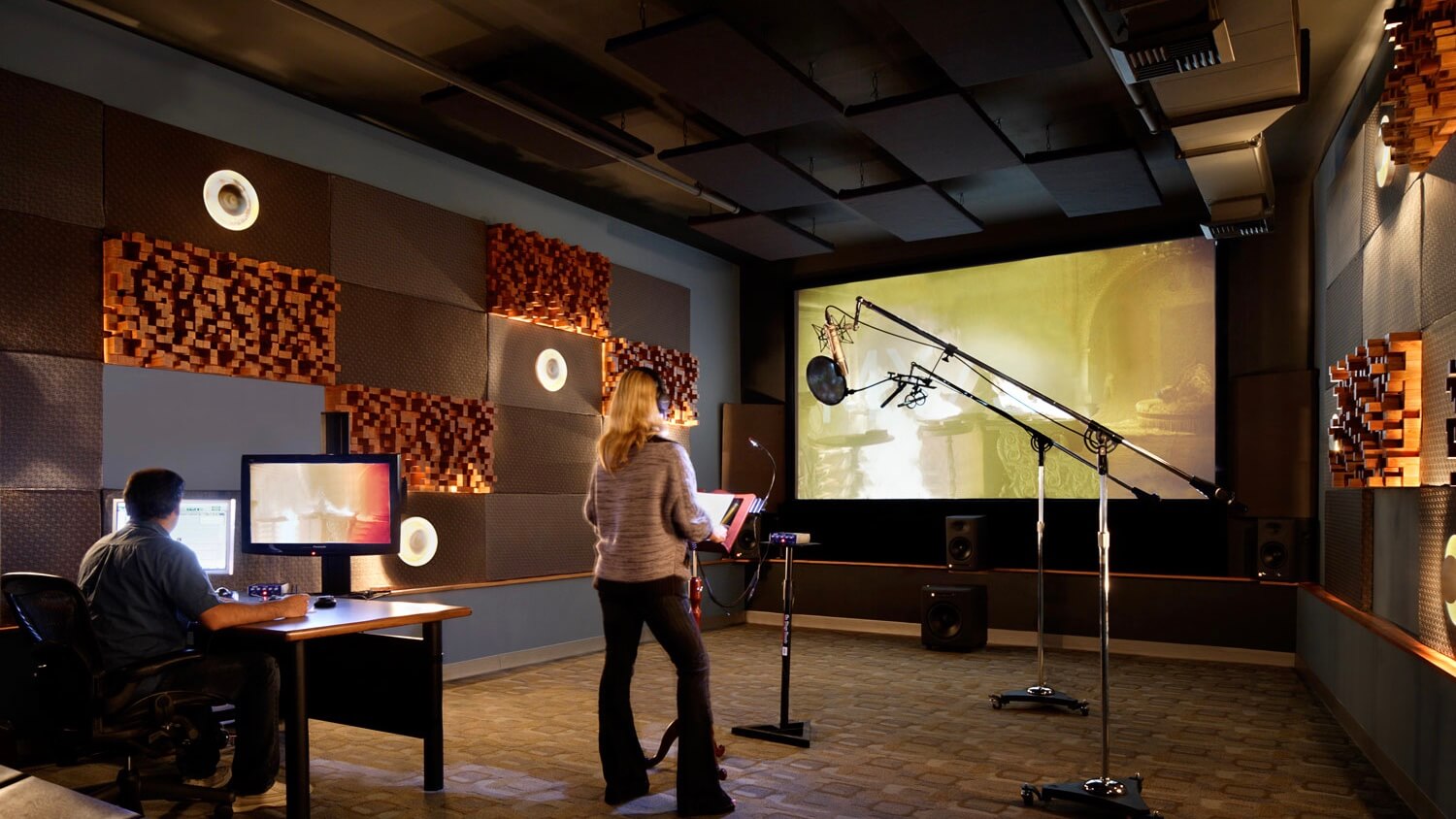 A film actor wearing headphones in a recording booth, reading lines into a large studio microphone while a dialogue editor watches through the control-room glass