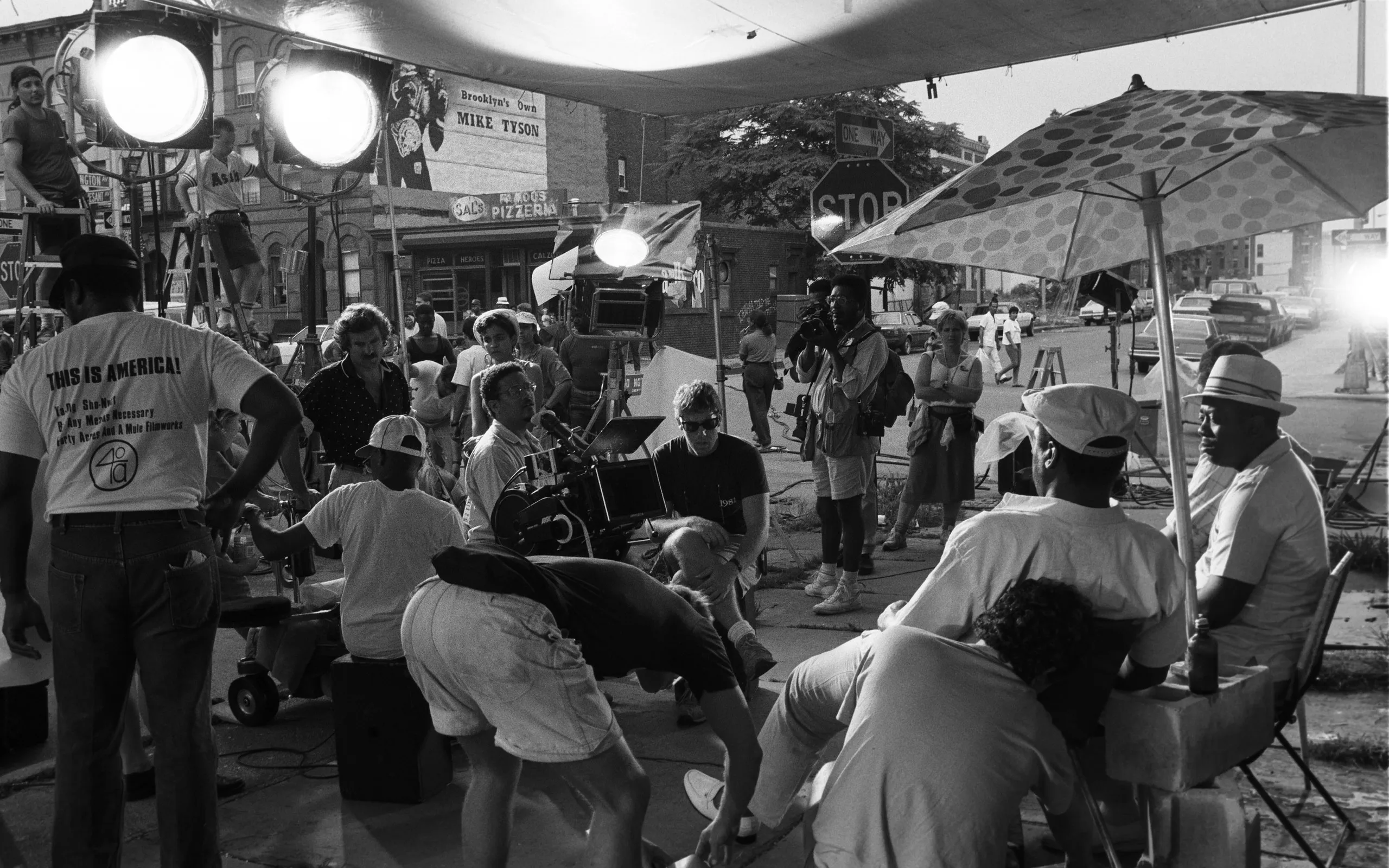 A film crew shooting on a city street with a camera on a tripod and lighting stands, while the area is partially cordoned off with cones