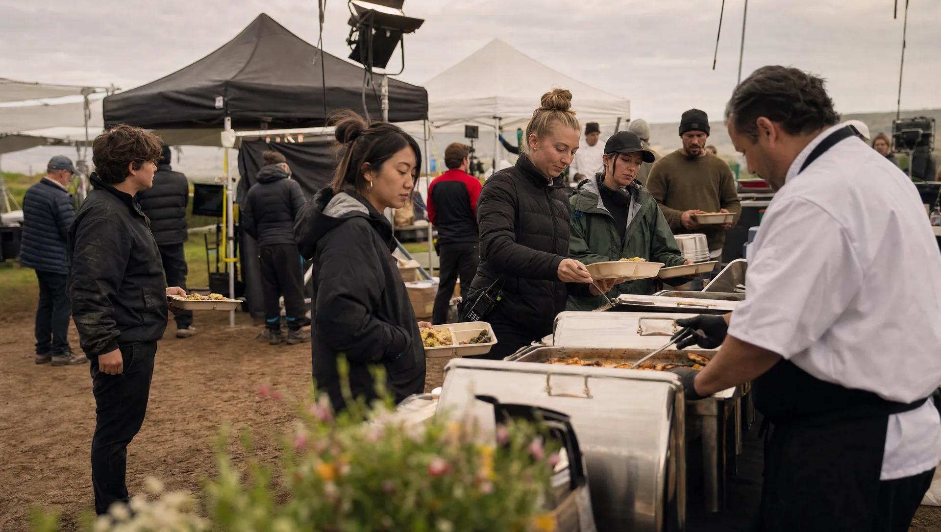 A film set meal table with hot trays, sandwiches, and drinks set up under a canopy near production trucks while crew members line up