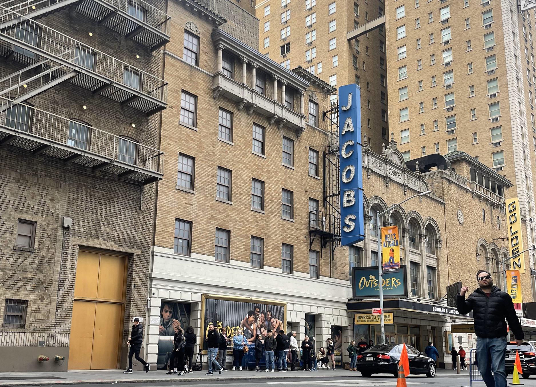 A line of patrons waiting at a Broadway theater box office at dusk, with marquee lights glowing above the entrance