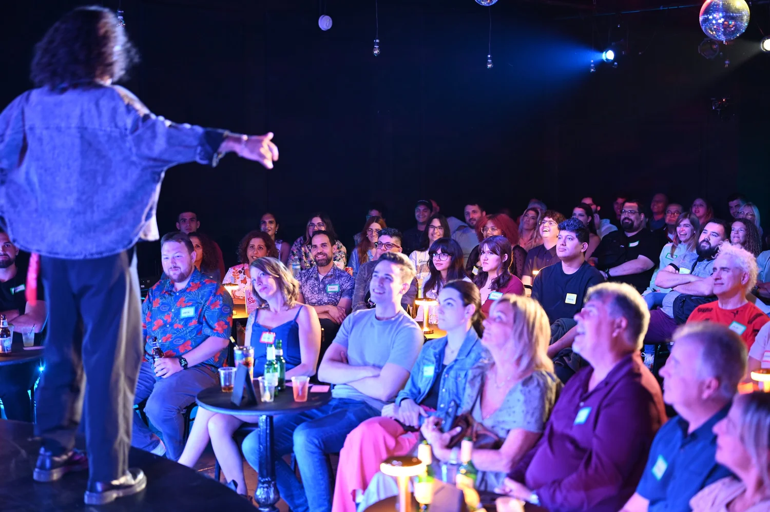 A lively stand-up comedy club with a comedian on stage under a spotlight and an audience seated at small tables