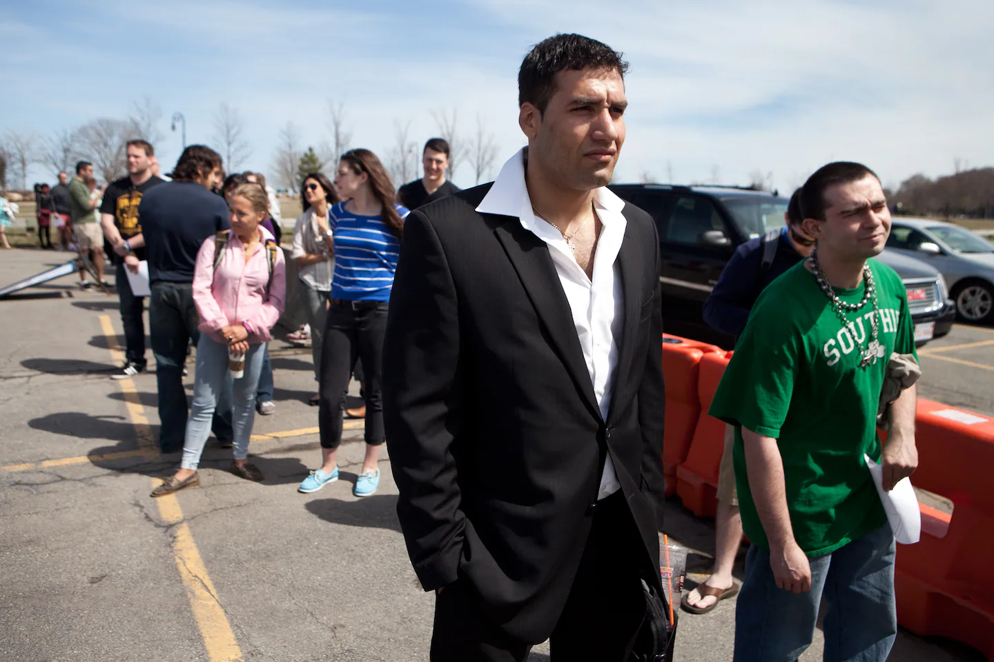 A long line of background actors waiting to check in outdoors near production signage, with a crew member holding a clipboard at a folding table