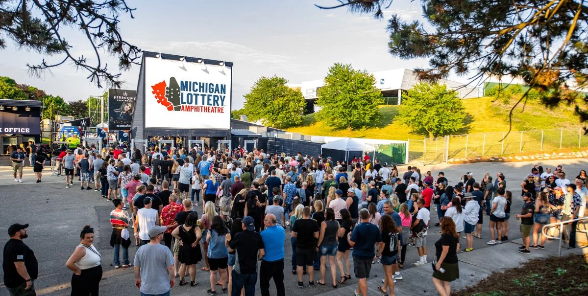 A long line of concertgoers waiting outside a venue entrance at dusk, security staff checking tickets near metal barricades