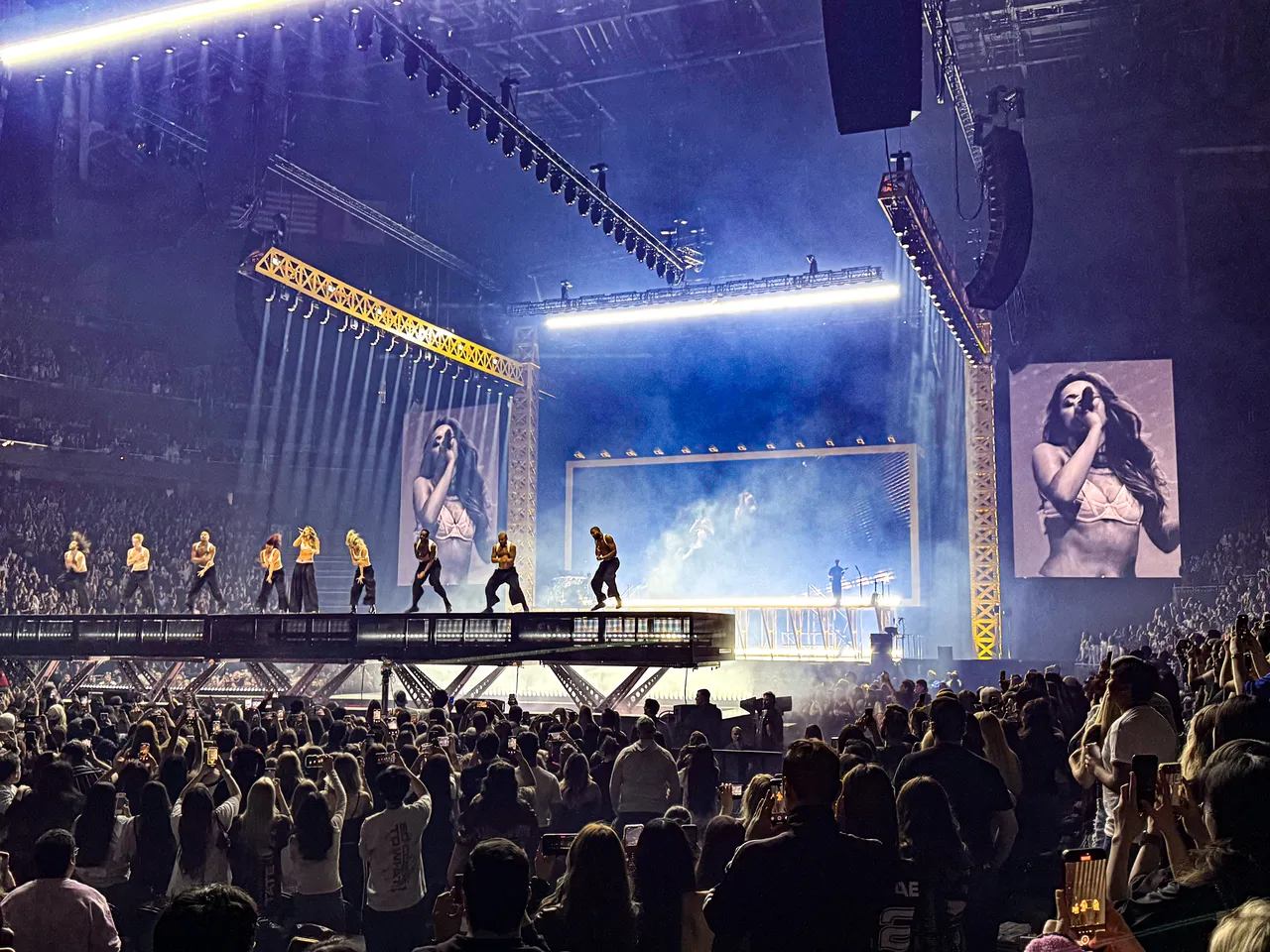 A packed concert pit at night with fans standing tightly together behind the metal barricade, the stage lights glowing in the background