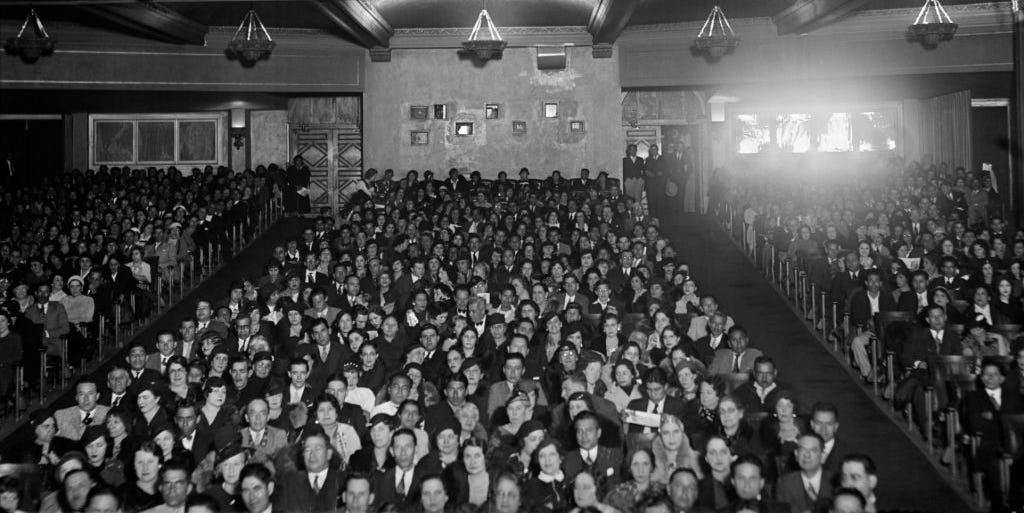 A packed movie theater auditorium during a large-format screening, with audience silhouettes facing a bright widescreen and soft aisle lights glowing