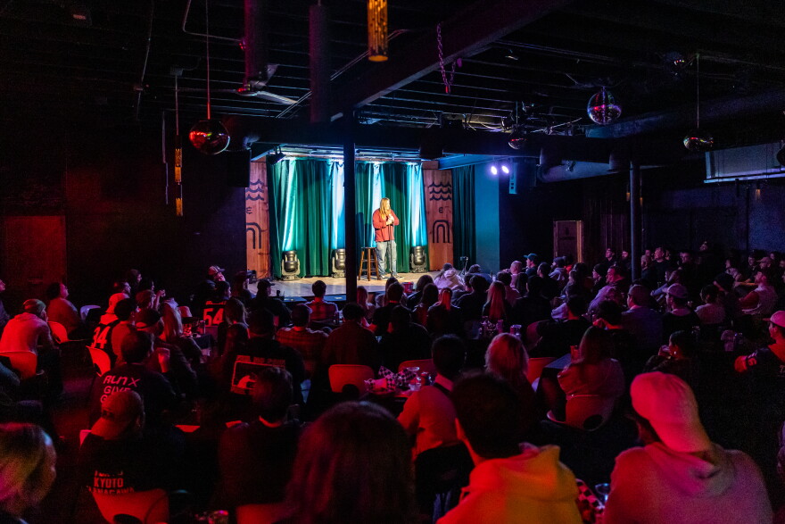 A photograph inside a dimly lit comedy club with a brick-wall stage and audience seated at small tables facing the microphone