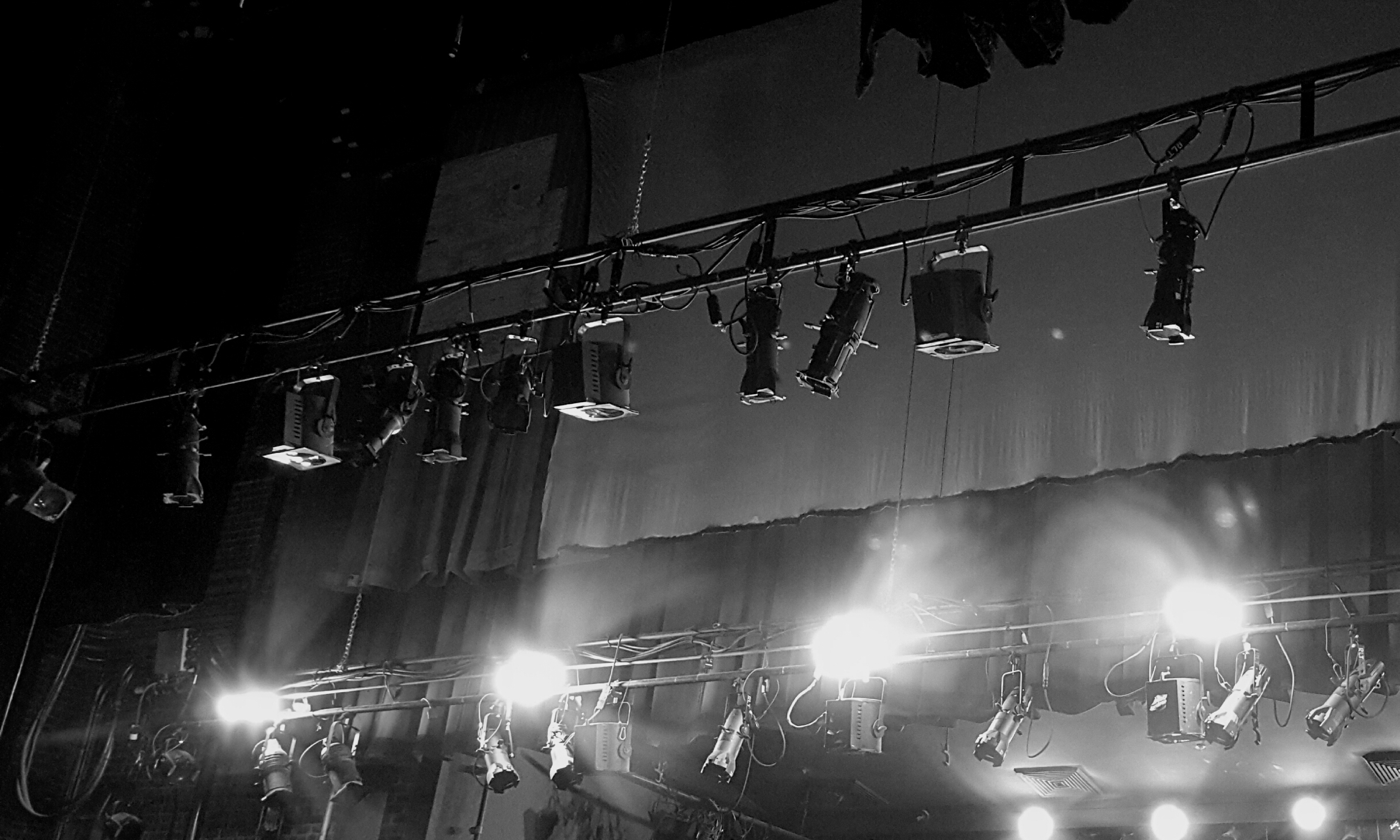 A row of ellipsoidal and Fresnel stage lights clamped to an overhead pipe in a theater, with safety cables visible