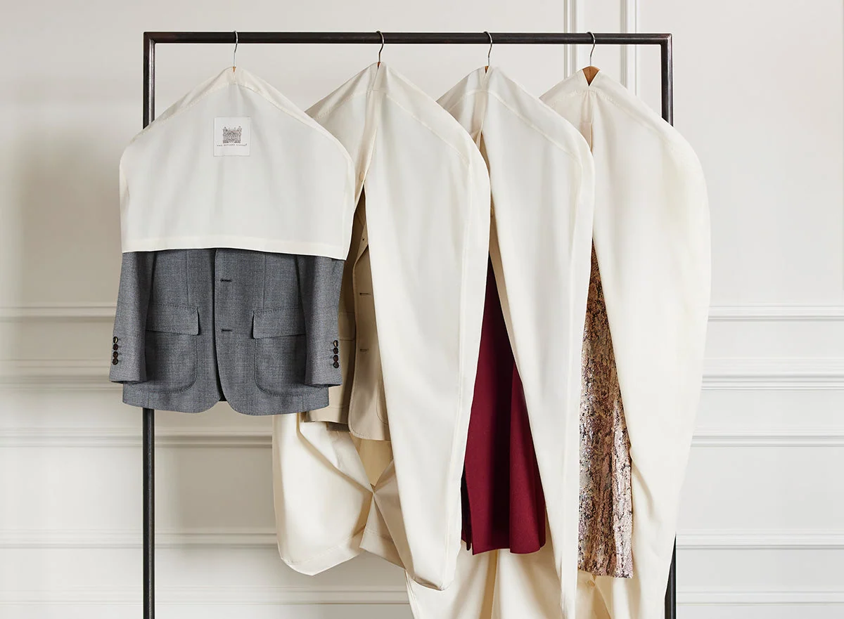 A row of garment bags labeled with paper tags hangs on a rolling rack in a production wardrobe area, with shoes lined neatly on the floor beneath