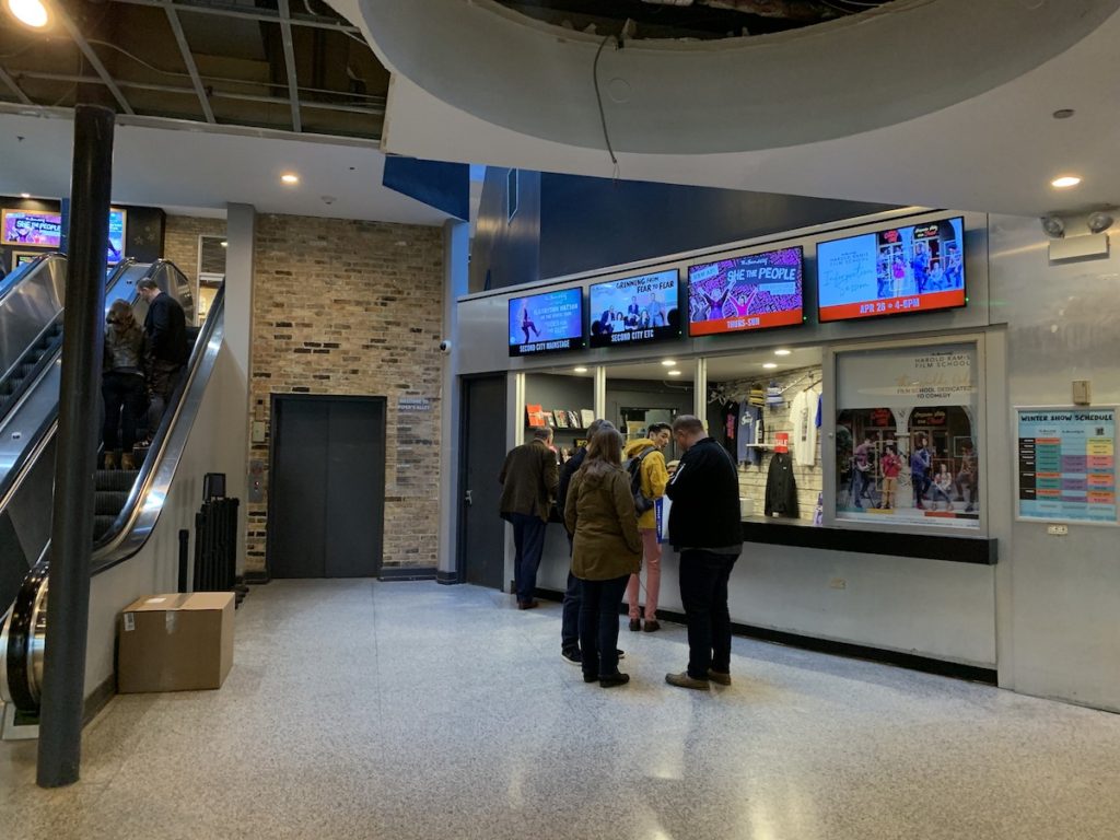 A small comedy club entrance with a ticket window and a line of patrons waiting outside at night