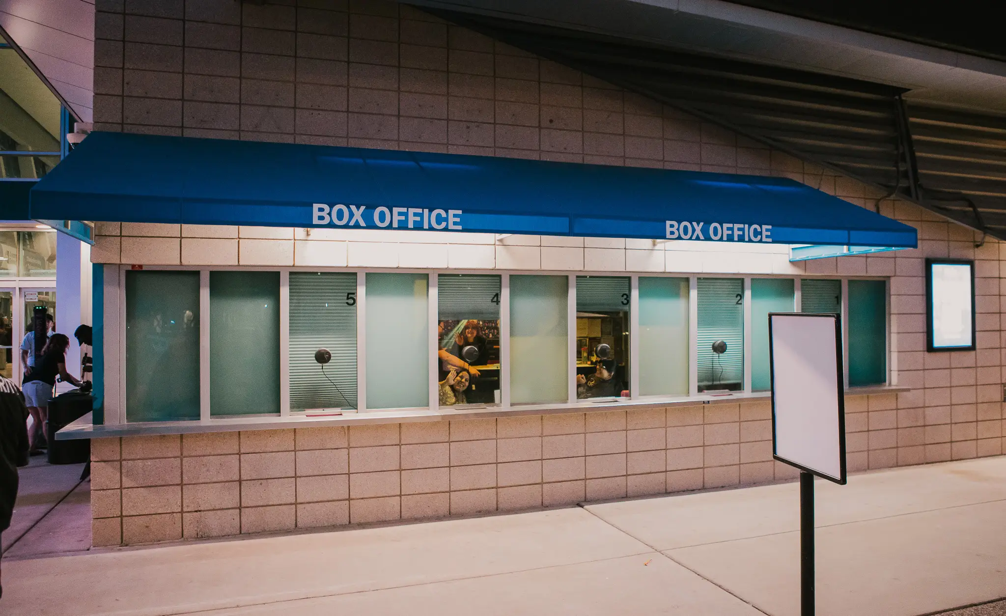 A small line of patrons waiting at a theater box office window on a city street in the early evening
