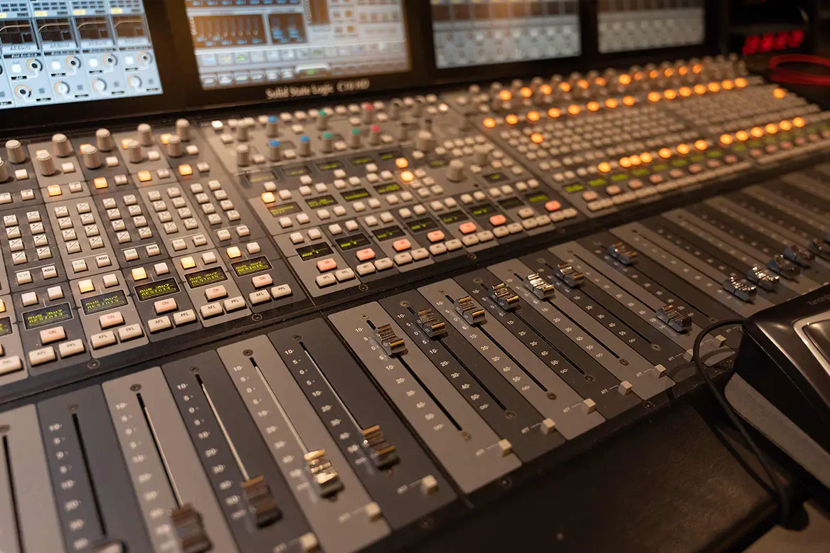 A sound engineer seated at a large mixing console in a post-production studio while a film scene plays on a screen in front of them