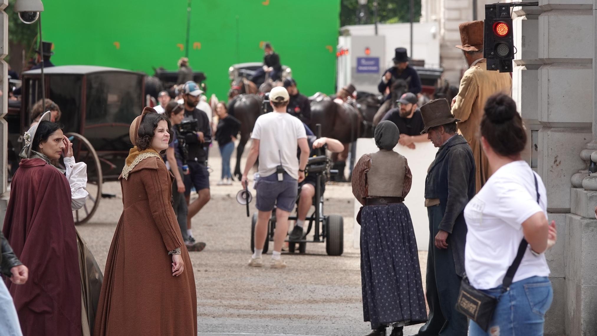 A stand-in actor marks a position on a film set while a camera operator frames the shot and lighting stands surround the scene