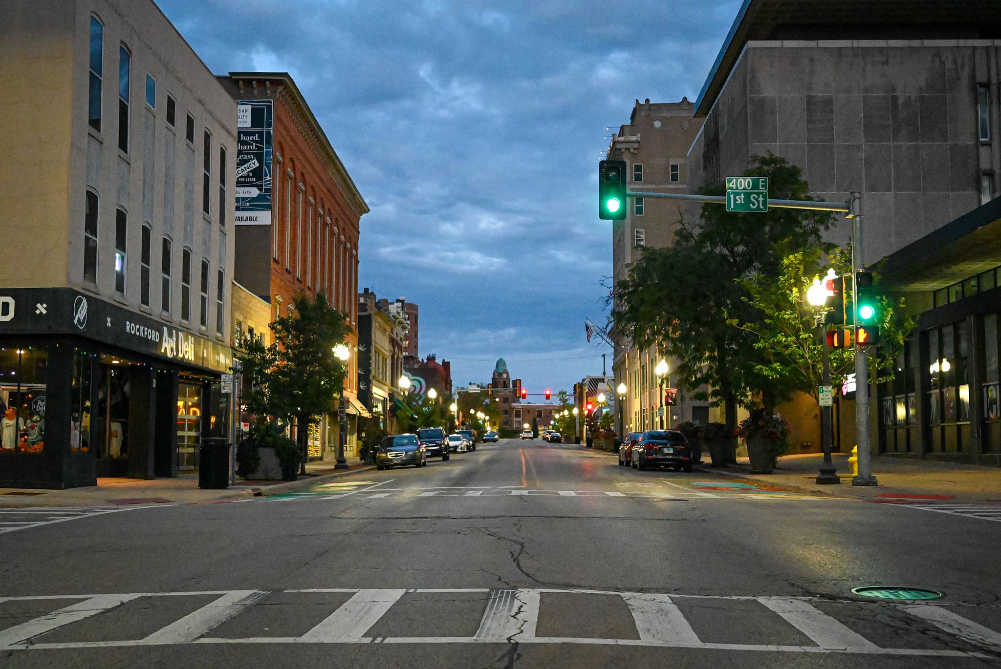 A street-level view of downtown Rockford, Illinois, with storefronts and cars parked along the curb.