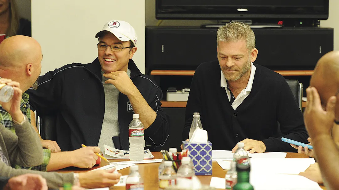 A television writers room with printed scripts, laptops, and coffee cups as producers and writers take notes while listening to actors read dialogue