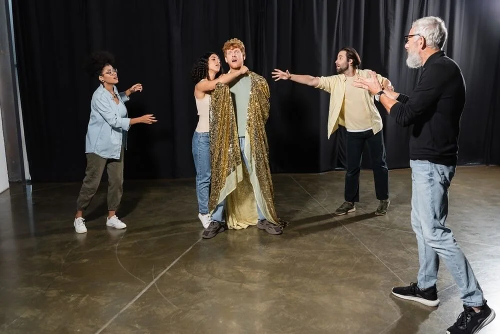 A theater director stands near the front row while actors rehearse positions on a lit stage with taped floor marks, during an early blocking rehearsal