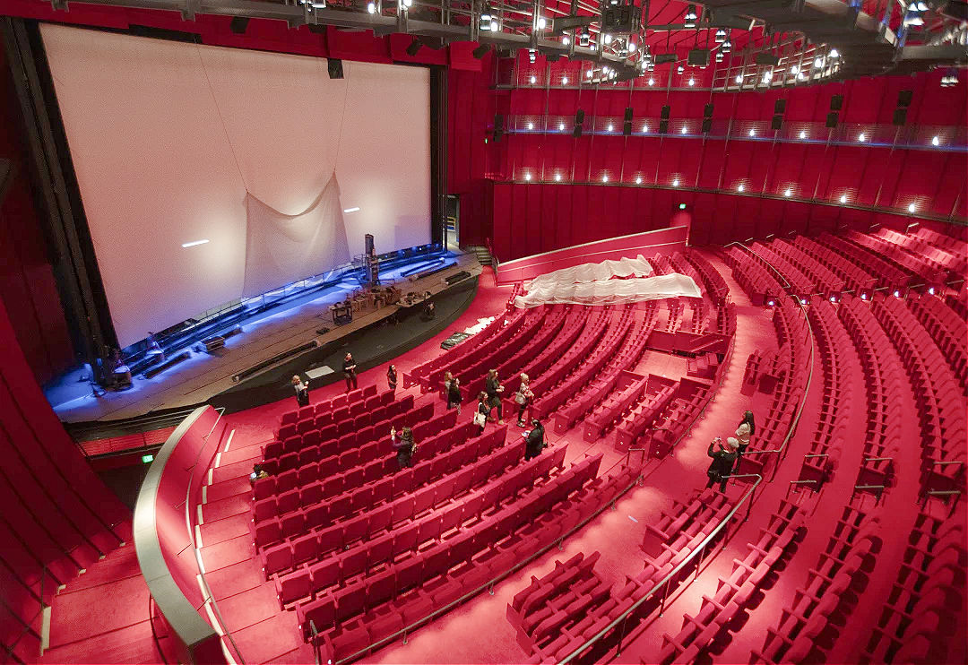 A theater stage in front of a large movie screen, with a host holding a microphone and audience members seated in dim light