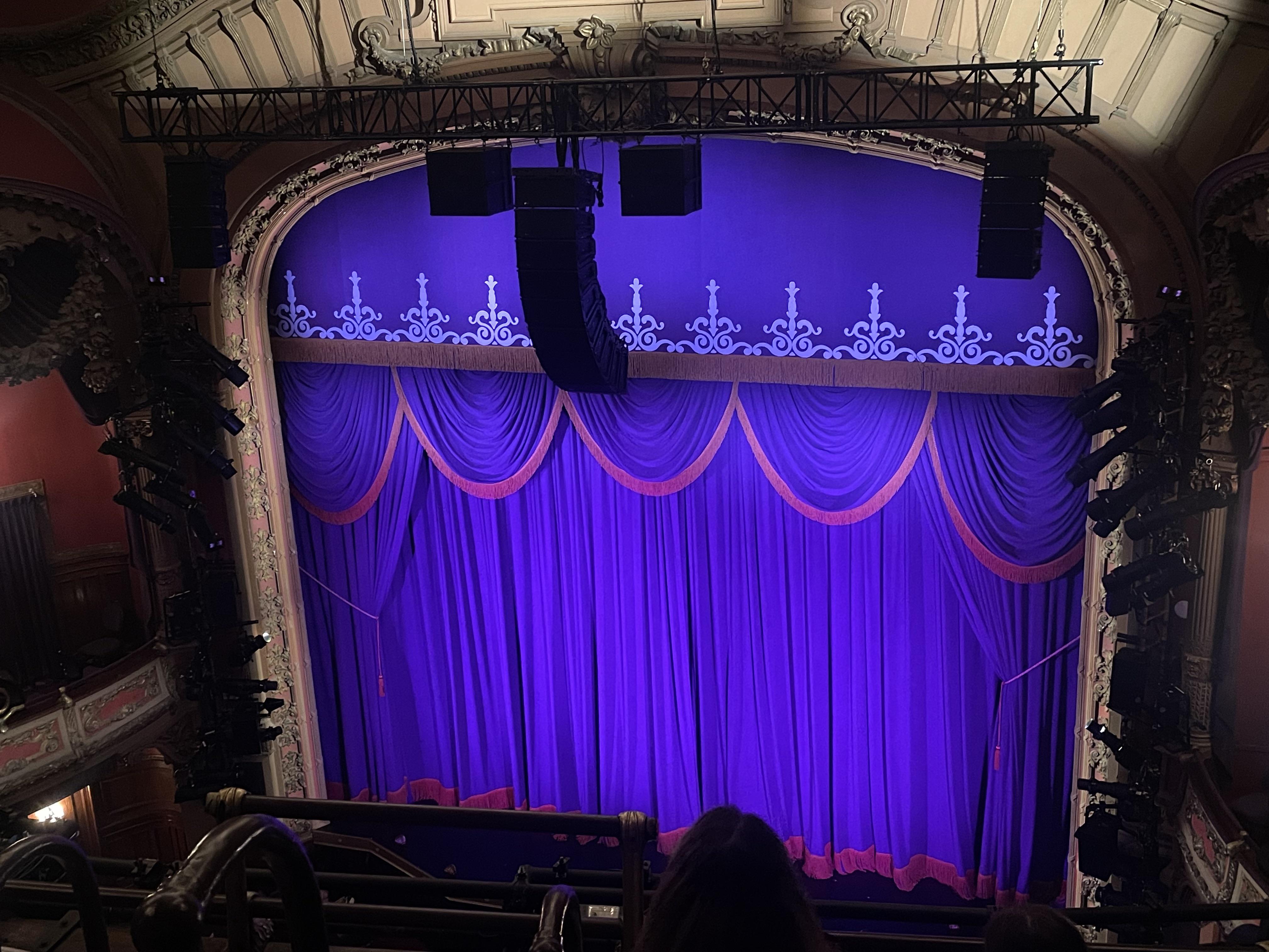 A view from a theater balcony looking toward a brightly lit stage with a single microphone stand set for a comedy show