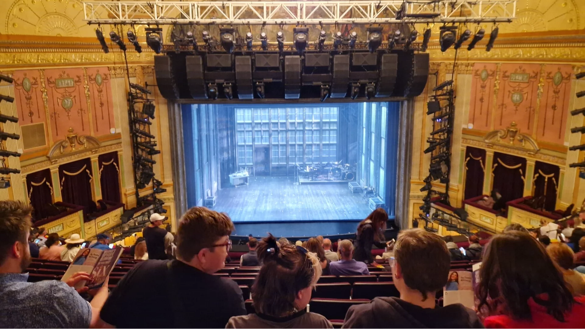 A view from the front mezzanine looking toward a brightly lit Broadway stage during a curtain call, with audience heads visible in the foreground