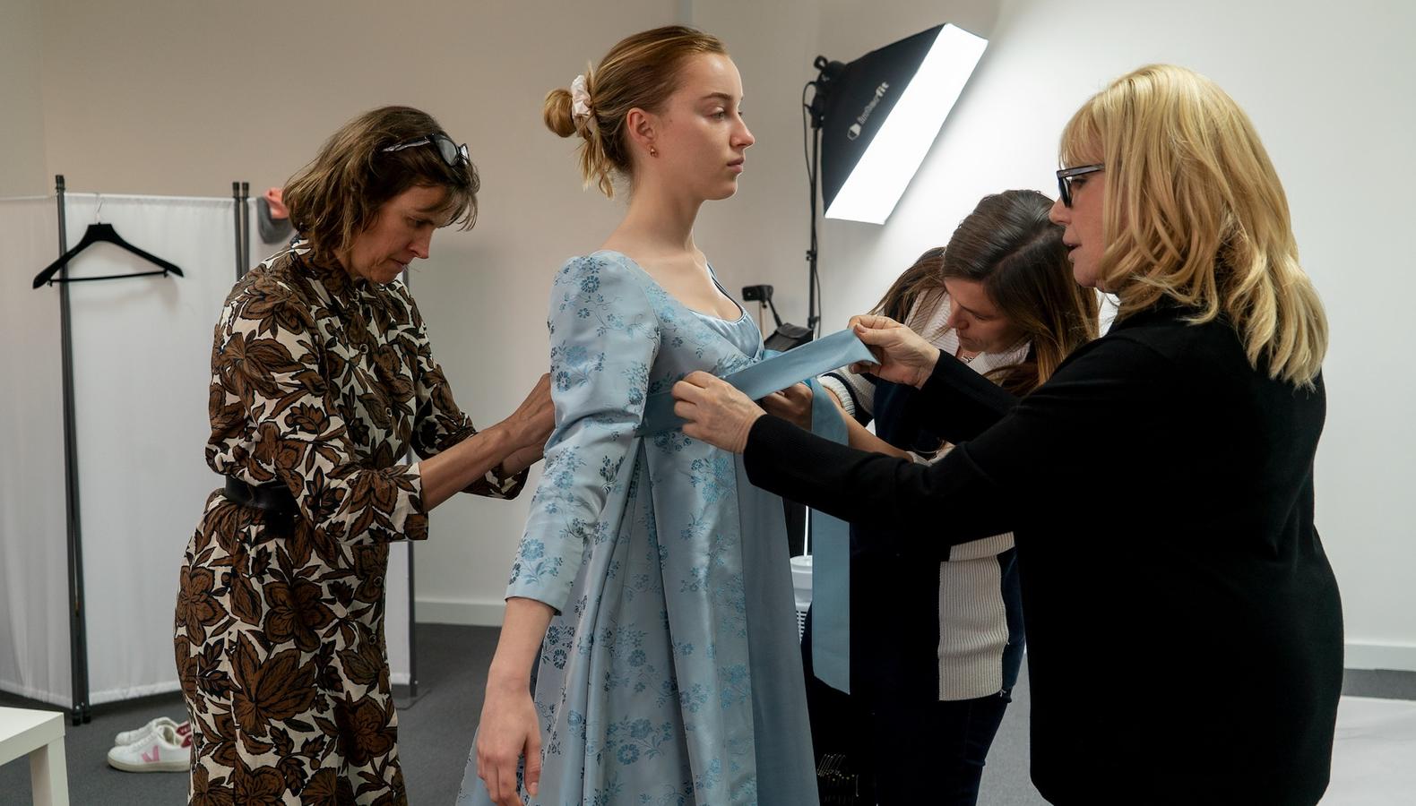 A wardrobe assistant adjusting the fit of a costume on a performer in a fitting area, with clothing racks and fabric swatches visible in the background