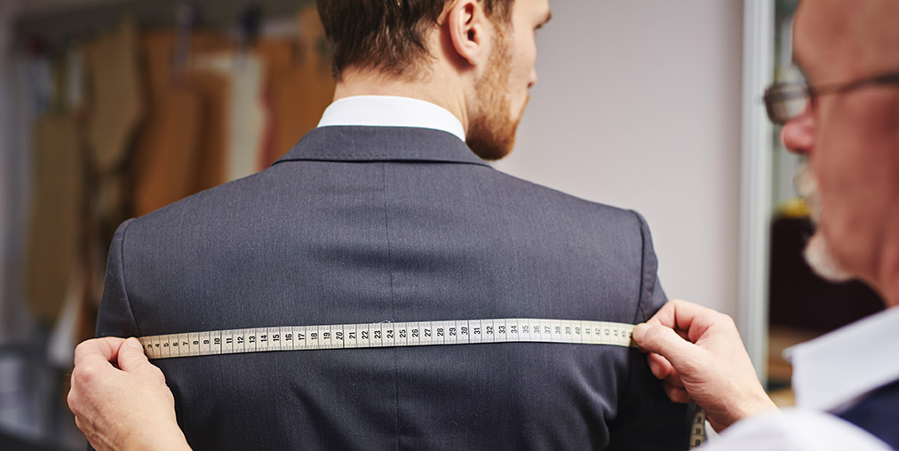 A wardrobe assistant measuring a background actor with a tape measure in a costume room filled with clothing racks