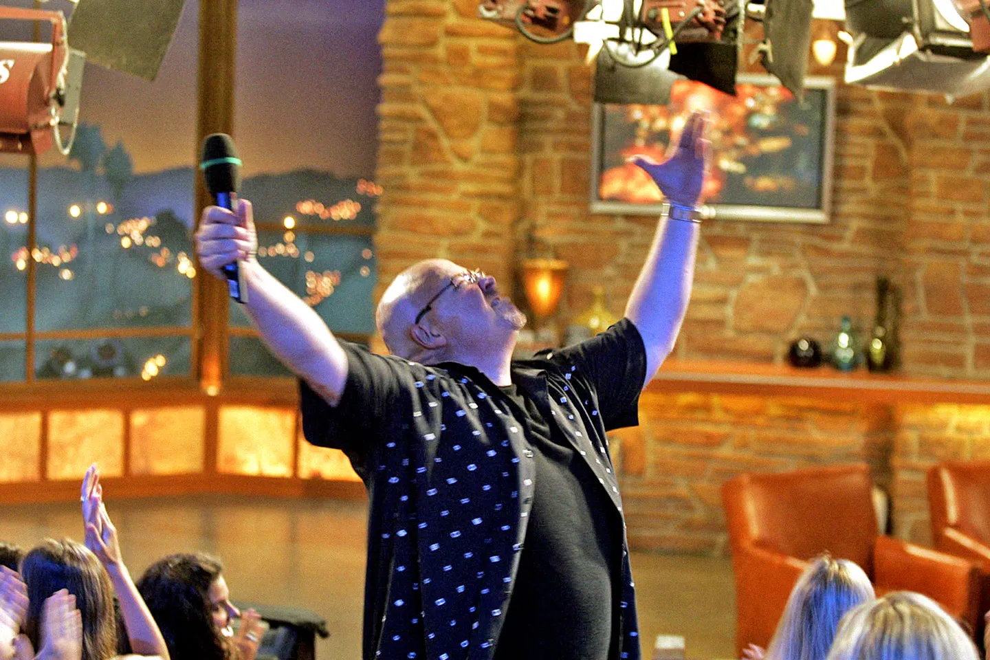 A warm-up performer on a small stage addressing a seated studio audience under bright production lights