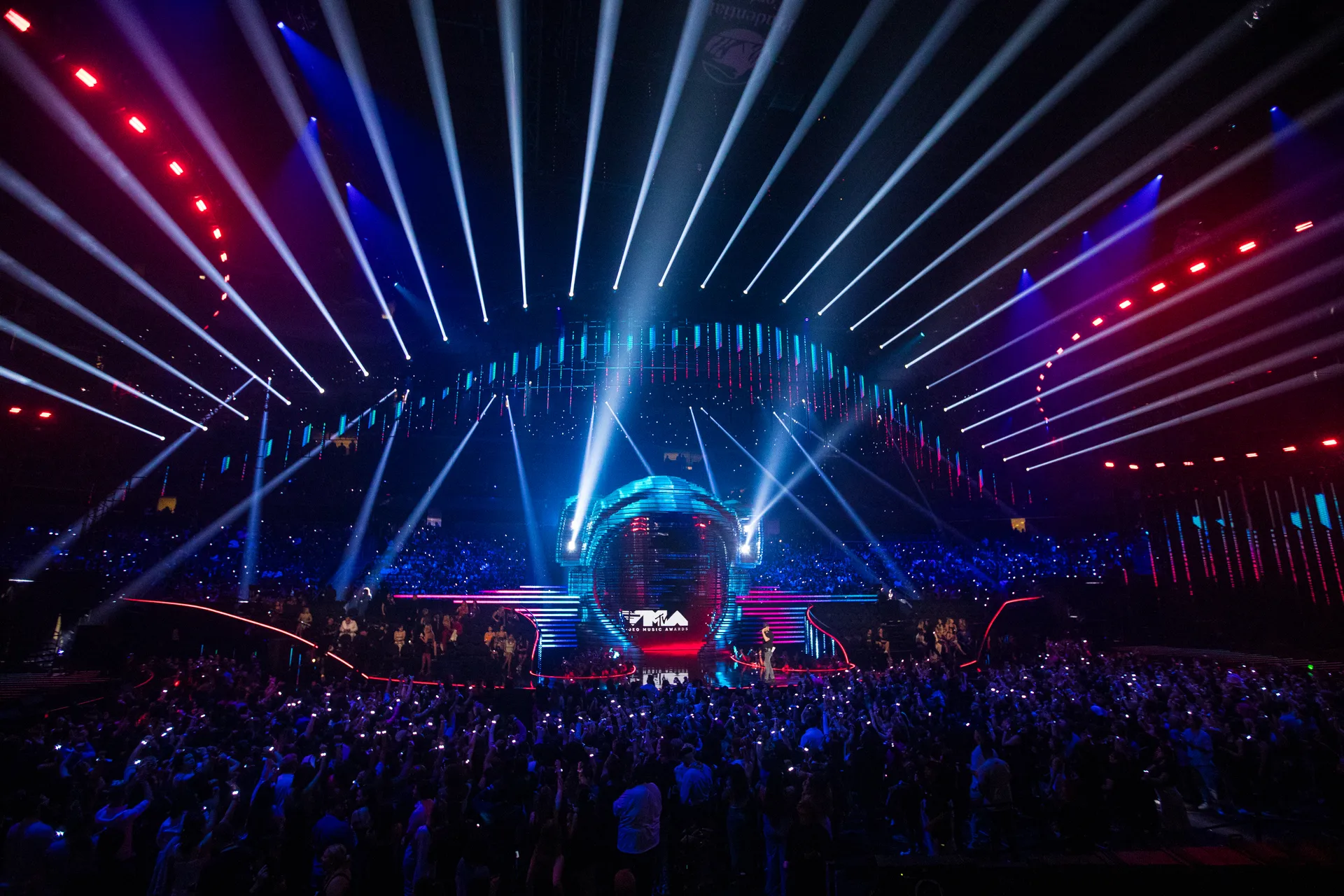 A wide shot of a VMAs stage during a live performance, with dramatic lighting beams, dancers in formation, and a large scenic set piece filling the background