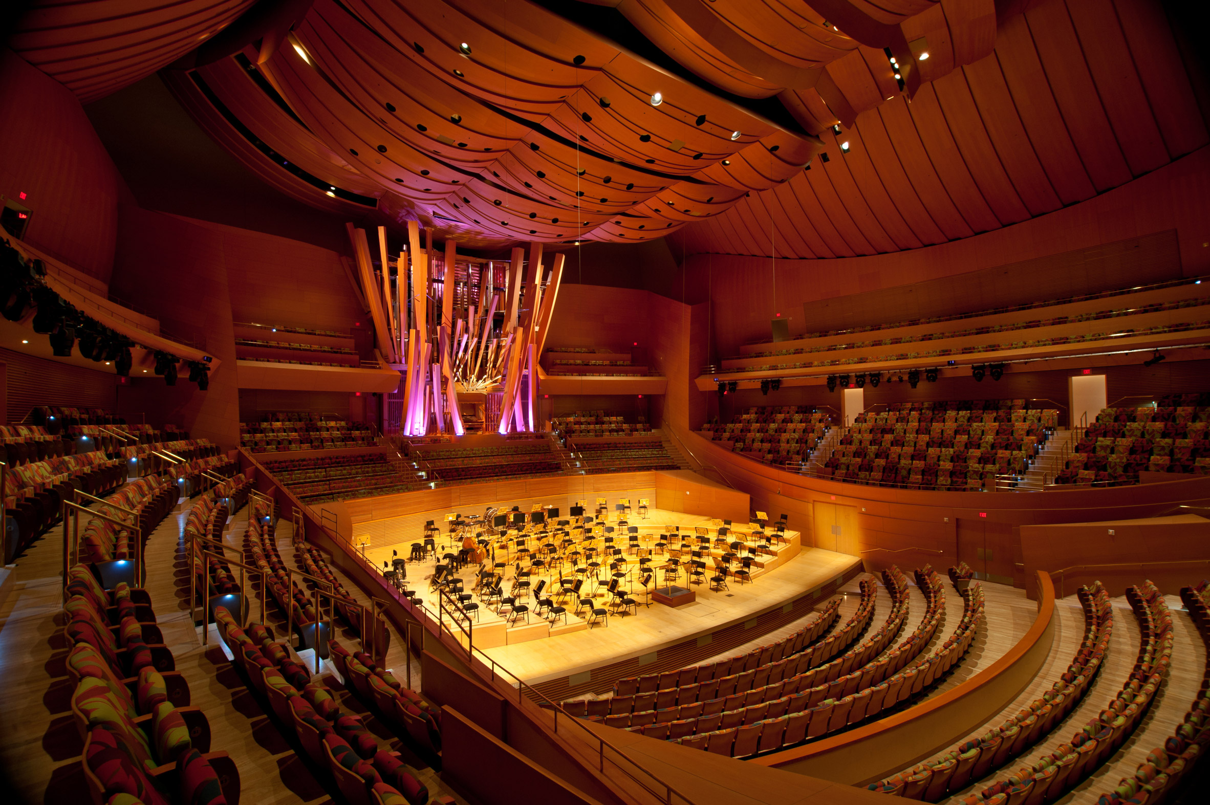 A wide view of a modern concert hall interior with a bright stage, terraced seating wrapping around the orchestra area, and balcony levels above