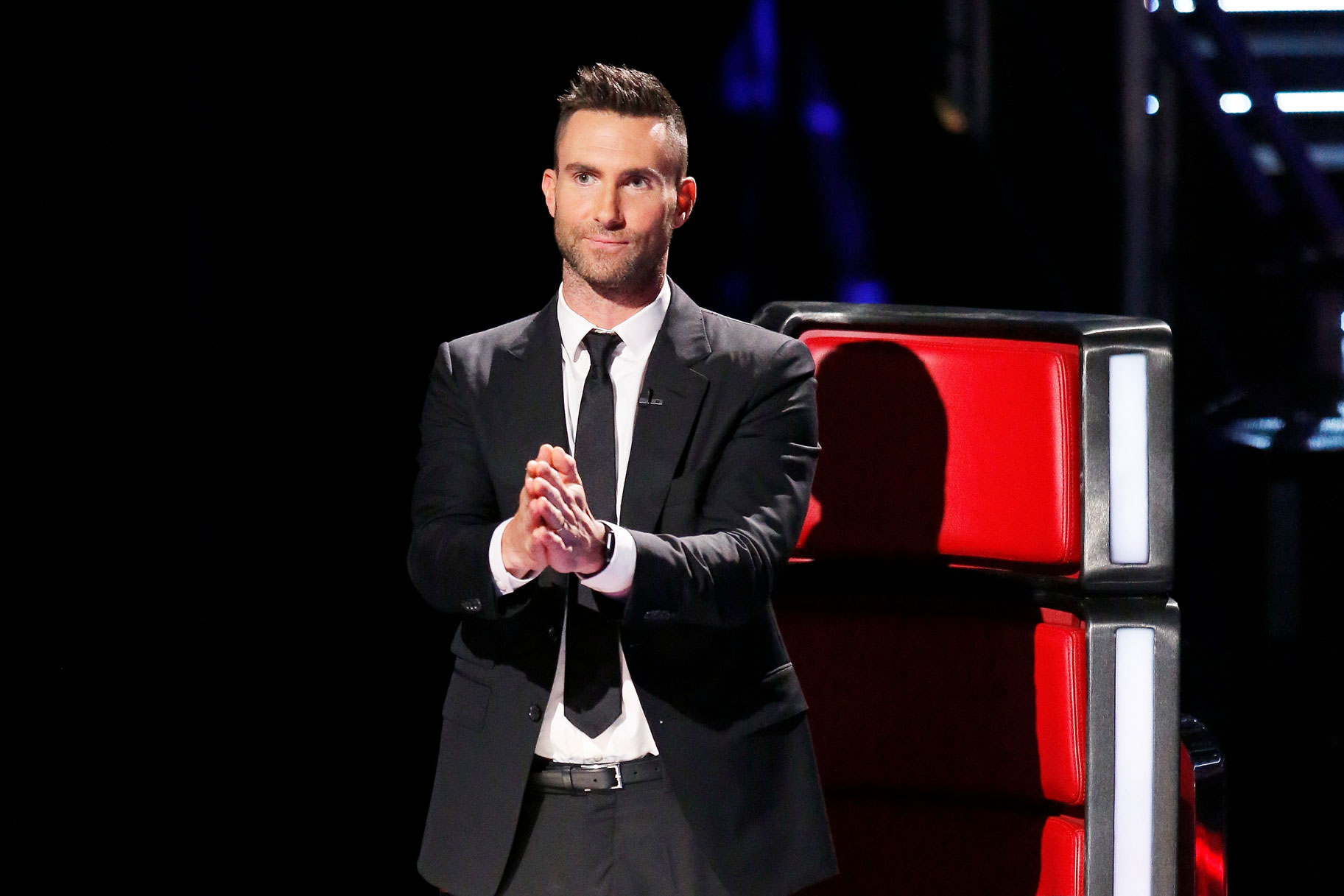 Adam Levine seated in a red coach chair on The Voice set, watching the stage with a focused expression