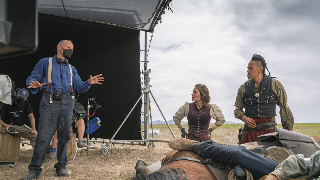 An assistant director holding a clipboard and walkie-talkie on a busy film set while crew members work in the background