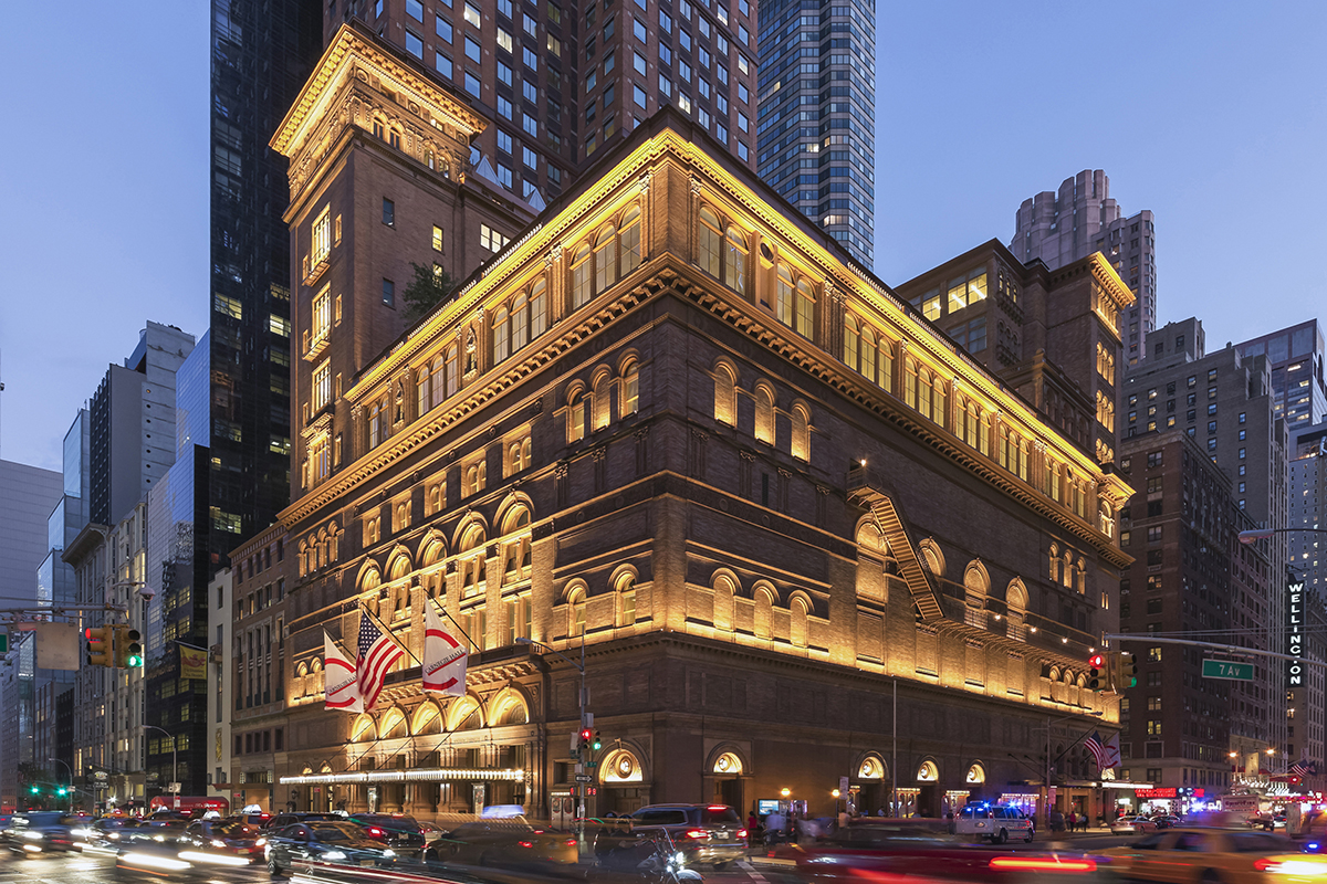 Carnegie Hall’s landmark facade in New York City at dusk, with pedestrians gathered near the entrance and warm lights glowing from the lobby