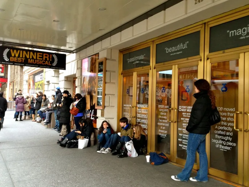 College students waiting in a line outside a New York City theater, wearing backpacks and checking their phones