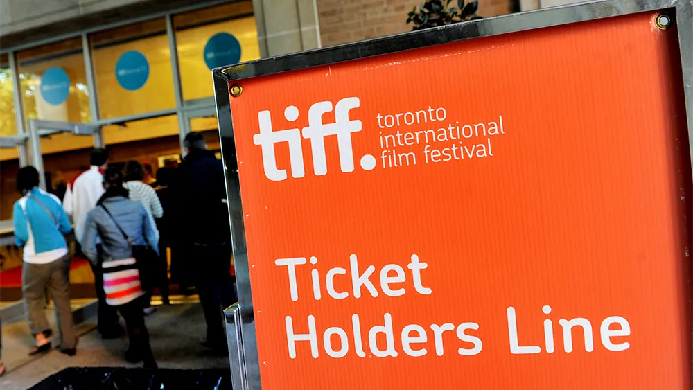 Festivalgoers waiting in a long line outside a downtown theater entrance, with marquee lights glowing at dusk and staff checking tickets