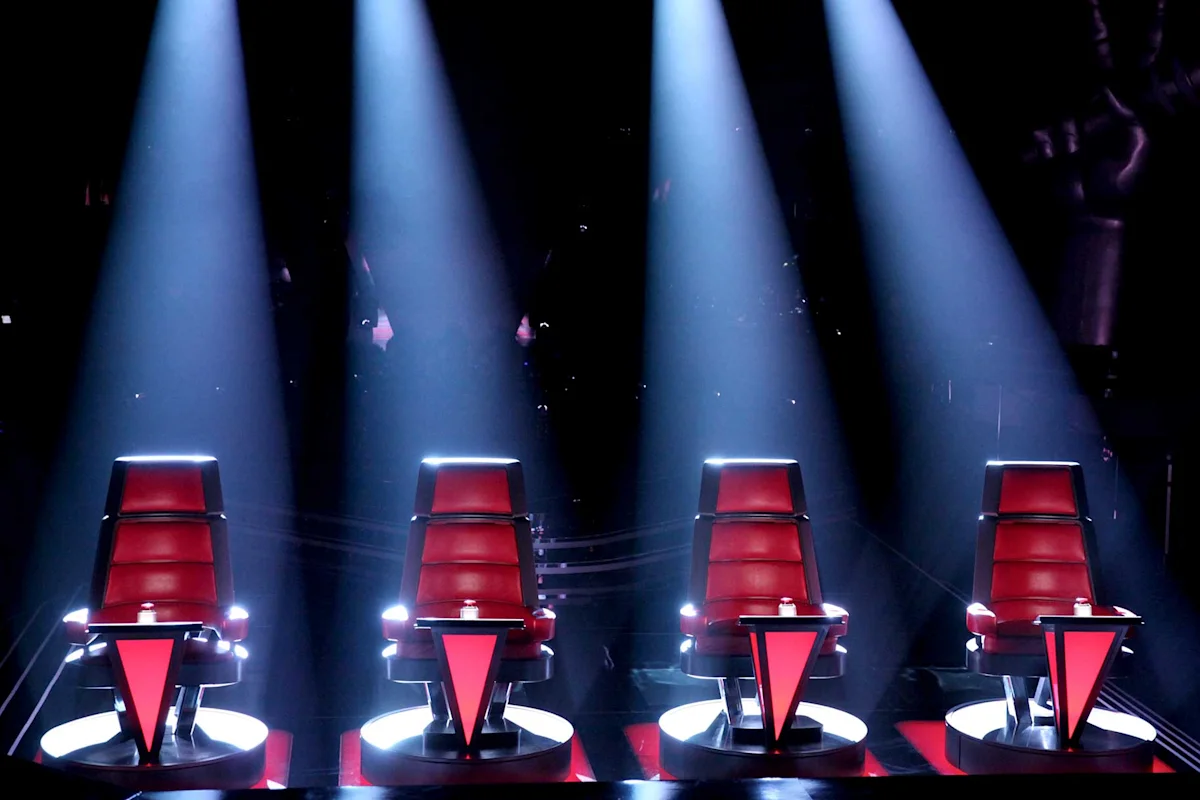 Four red swiveling coach chairs facing away from a brightly lit stage on a television singing competition set