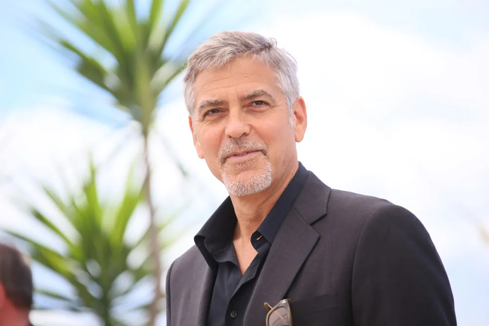George Clooney at a 2020-era public appearance, standing against a neutral backdrop and smiling slightly.