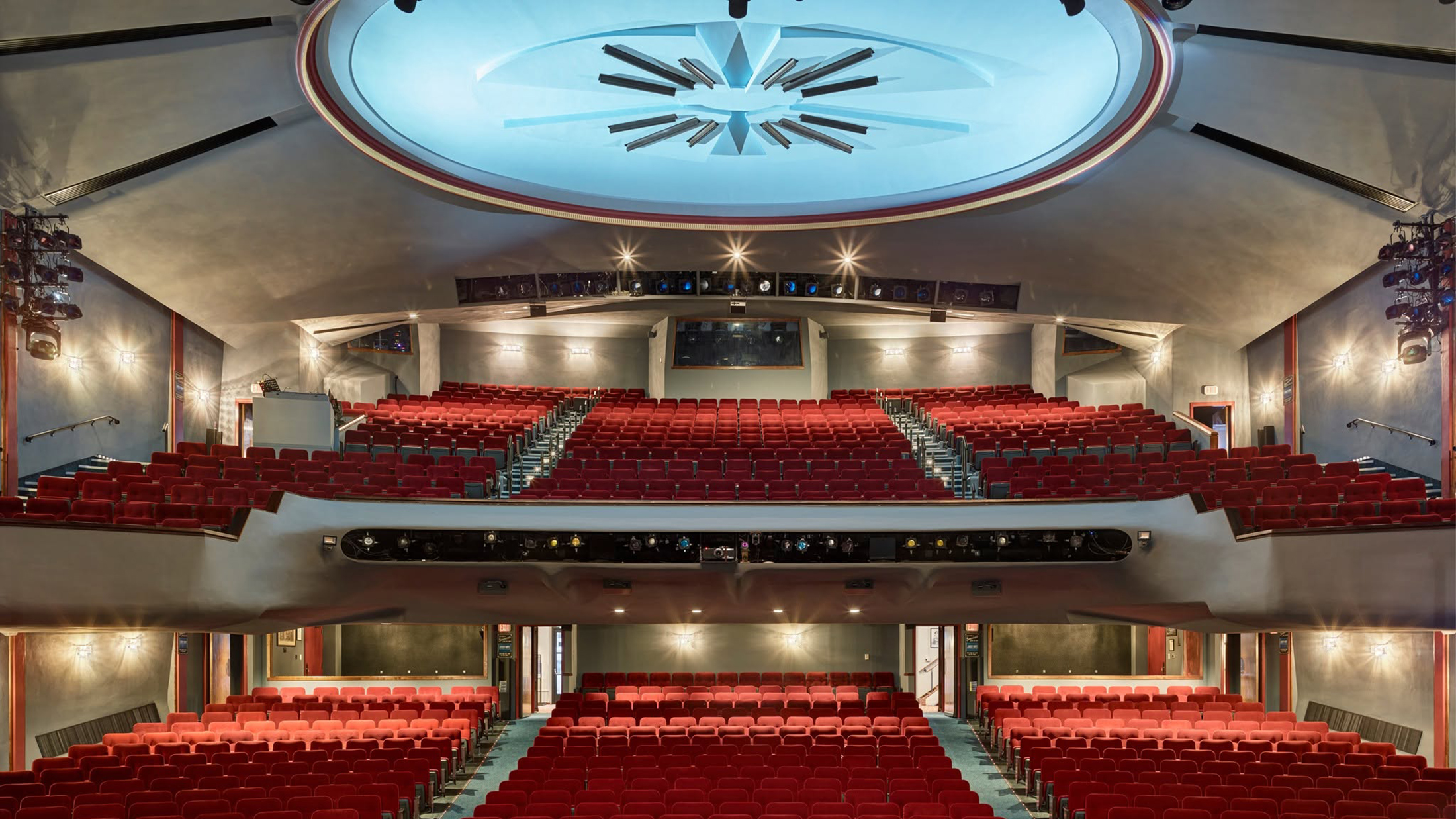 Interior of a small regional theater with a modest stage and closely spaced audience seating, lit warmly before the show begins