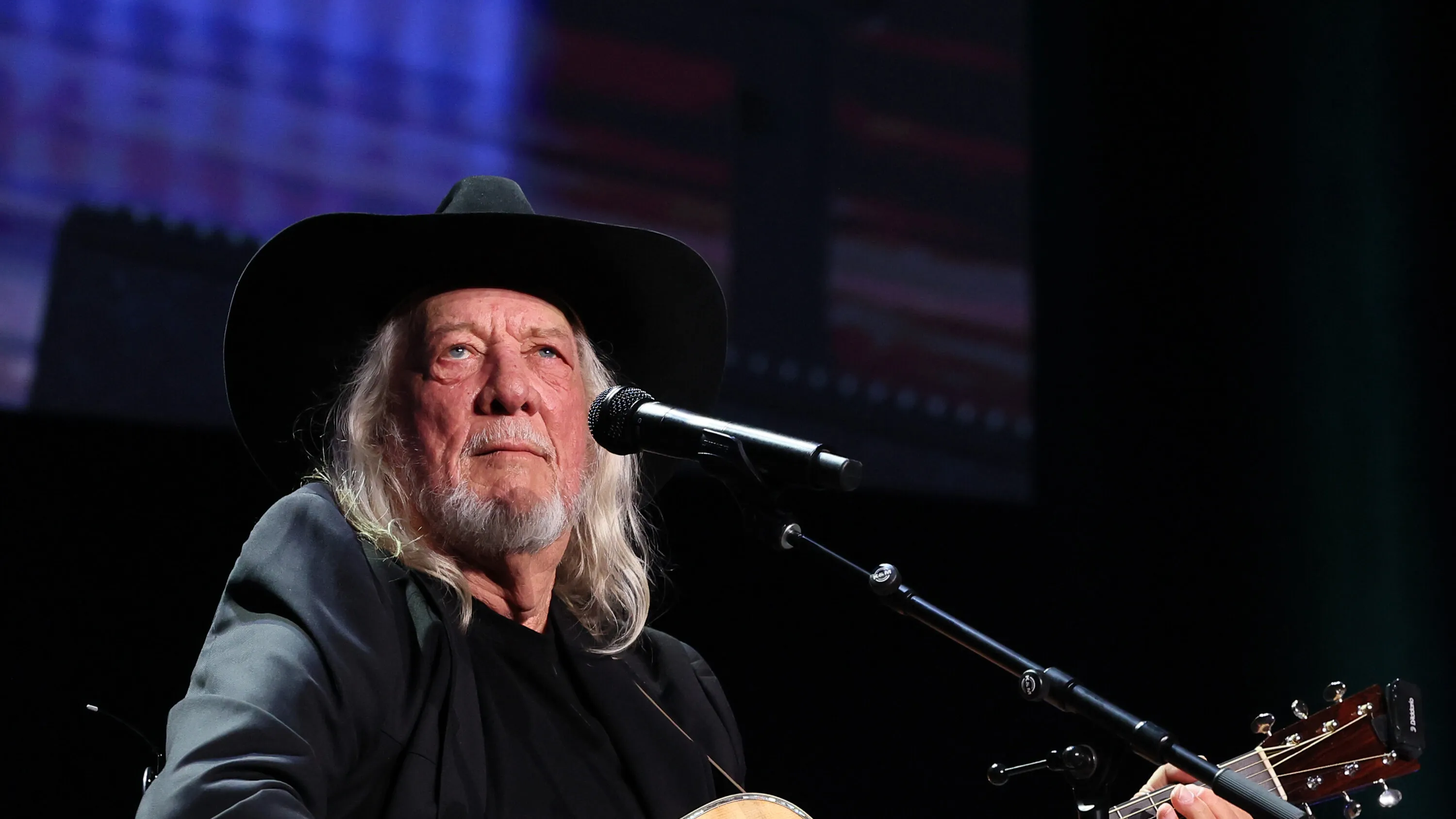 John Anderson sings into a microphone onstage with band members behind him and stage lights shining overhead.
