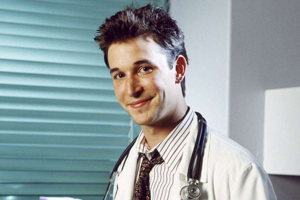 Noah Wyle seated for an interview, wearing a simple jacket, hands lightly clasped, looking attentive under soft studio lighting.